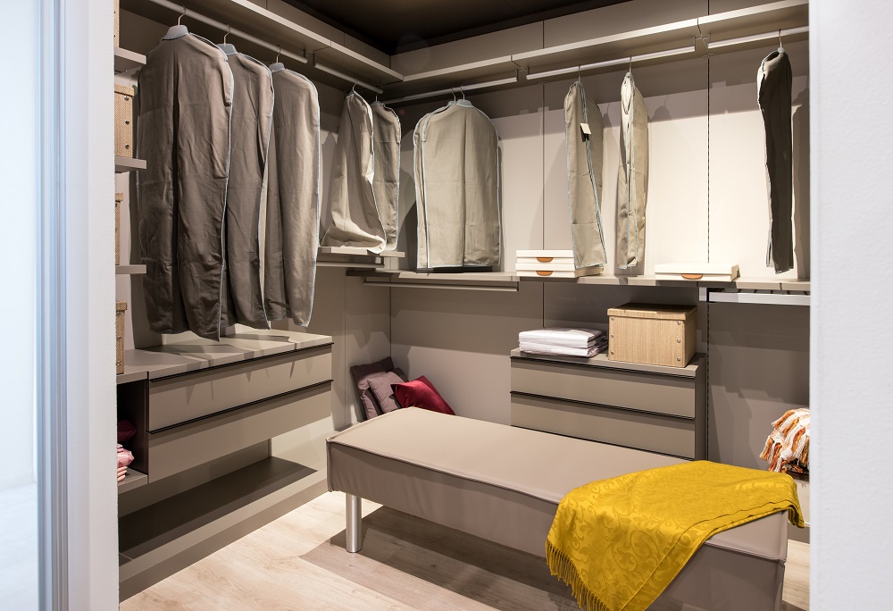 Interior of a walk in closet with hanging clothes and built in drawer units and seating in a luxury modern home